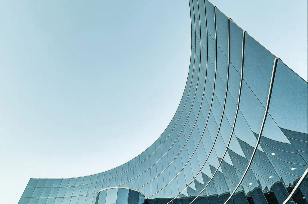 This image shows a striking example of modern architecture, featuring the sleek, curved facade of a glass building that mirrors the clear blue sky. The building's reflective surface creates a fluid, wave-like appearance, blending the boundaries between structure and sky.