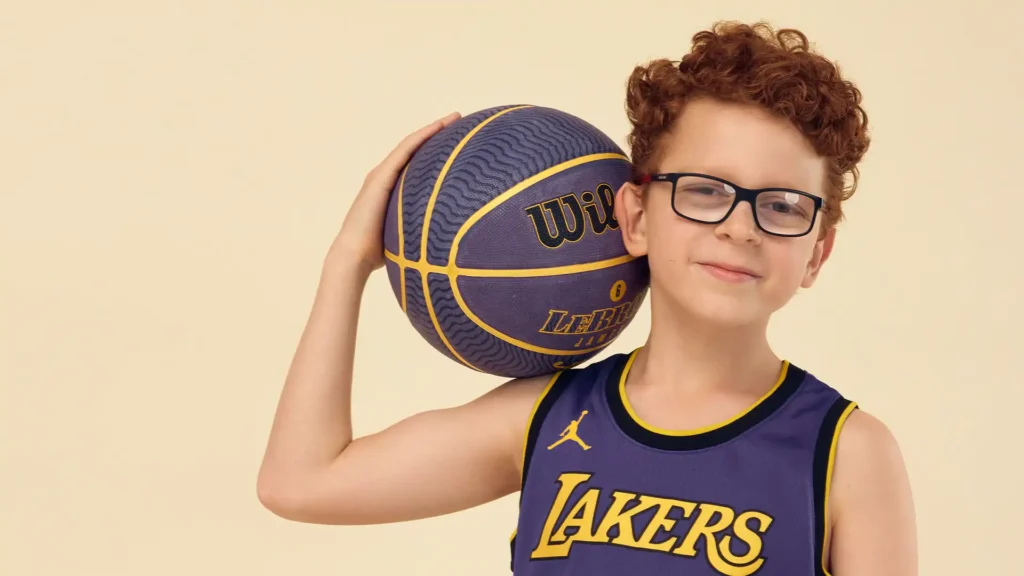 A smiling young boy with curly hair, wearing glasses and a Los Angeles Lakers basketball jersey, holds a basketball on his shoulder against a light beige background. He looks confident and playful, ready for a game of basketball.A smiling young boy with curly hair, wearing glasses and a Los Angeles Lakers basketball jersey, holds a basketball on his shoulder against a light beige background. He looks confident and playful, ready for a game of basketball.