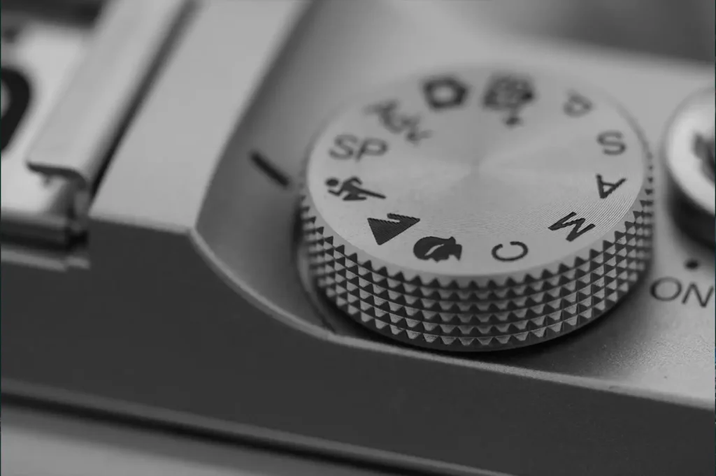 This image is a black and white close-up of a camera's mode dial, with a shallow depth of field emphasizing the textured grip and iconic camera settings symbols. The monochromatic tones highlight the intricate details and classic design elements of the camera, evoking a sense of nostalgia and precision associated with photography.