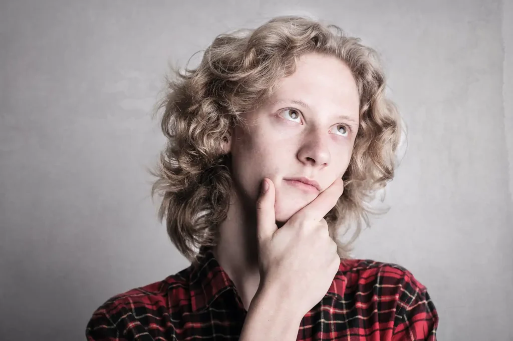 A contemplative teen model with curly blond hair, wearing a red and black checkered shirt, is gazing thoughtfully upwards to the side. The person's chin rests on their hand, suggesting a moment of deep thought or decision-making. The background is a neutral grey, drawing focus to the subject's introspective expression.