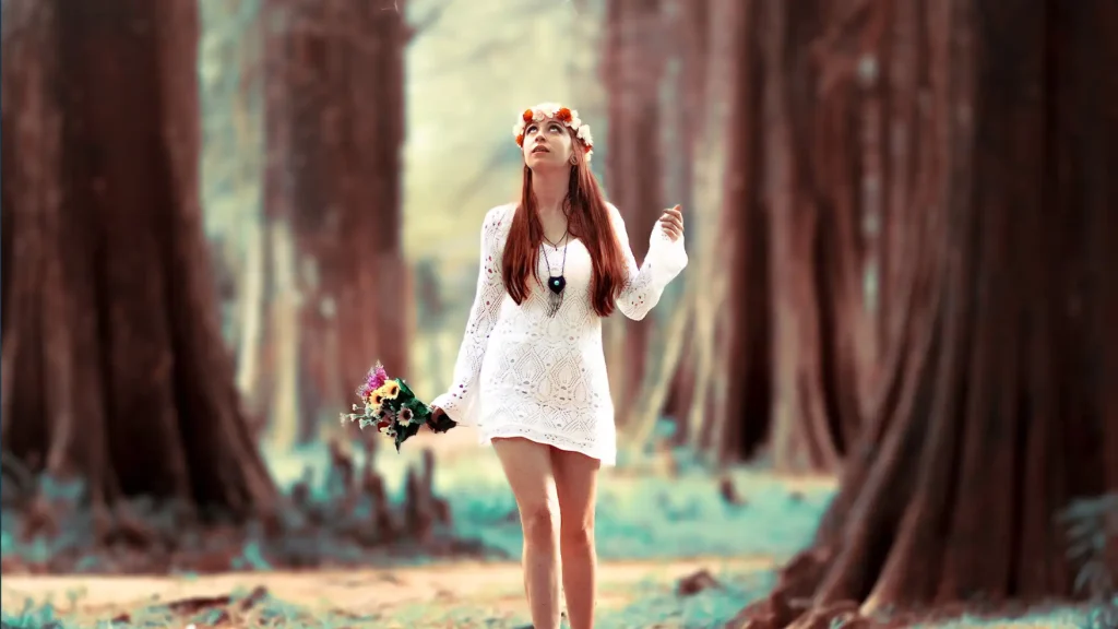 A teen model with long hair wearing a white lace dress and a floral headband stands in a serene forest. She is holding a bouquet of flowers and looking up, possibly towards the sky, surrounded by tall, majestic trees with a soft, dreamy ambiance.