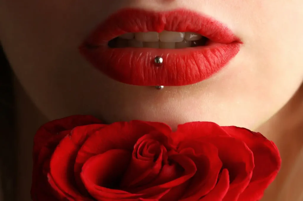 A close-up image showing the lower half of a person's face with striking red lipstick matching the vibrant red of a rose positioned directly below the lips. The person has a labret piercing below their lower lip, and the photo focuses sharply on the sensual curves of the lips and the intricate petals of the rose, creating a strong visual association of color and texture.