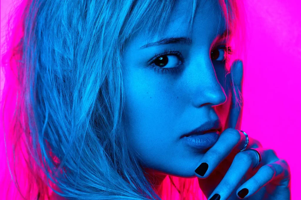 A close-up image featuring a teen model in a neon blue and pink light setting, creating a vivid and dynamic visual effect. The subject's contemplative expression and direct gaze are highlighted by the contrasting colors, with details such as their dark nail polish and subtle jewelry adding depth to the portrait. The image exudes a modern and edgy vibe, indicative of contemporary portrait photography.