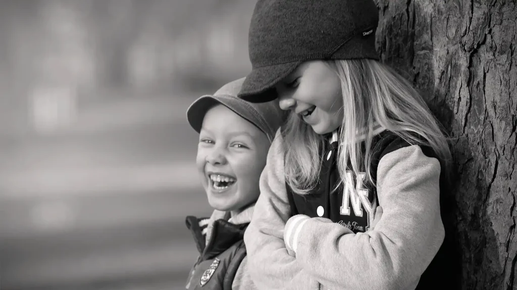In a heartwarming black and white photograph, two children share a genuine moment of joy and laughter. Leaning against a tree, the boy on the left, wearing a cap and a joyful smile, appears to be sharing a funny moment with the girl on the right, who is also wearing a cap and laughing heartily. Their close proximity and comfortable body language suggest a strong bond, possibly siblings or close friends, delighting in each other's company in a park or outdoor setting. The candid shot captures the innocence and happiness of childhood.