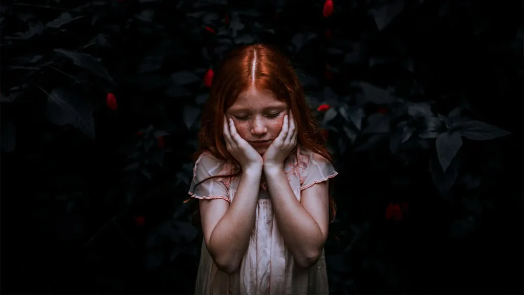 A contemplative young girl with red hair and freckles stands against a dark backdrop of leaves with sparse red berries. Her eyes are closed, and her face is gently cupped in her hands, suggesting a moment of introspection or sadness. The contrast between her light skin and the dark surroundings creates a dramatic and emotive scene.