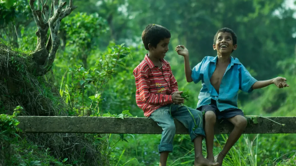 Two joyful young boys are sitting on a rustic concrete beam in a lush green setting, possibly in a rural area. The boy on the left, wearing a red-striped shirt and denim shorts, is watching his companion, who is animatedly telling a story, indicated by his expressive hand gestures and wide smile. The boy on the right, in an unbuttoned blue shirt and shorts, exudes excitement and happiness, his feet bare and his laughter possibly echoing through the serene environment. This image captures a moment of pure, carefree childhood friendship.