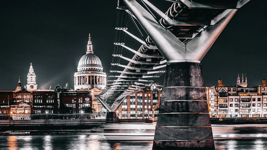 A night-time scene captures the iconic St. Paul's Cathedral and the modern Millennium Bridge over the Thames River in London, with city lights reflecting off the water, juxtaposing historic architecture with contemporary design.