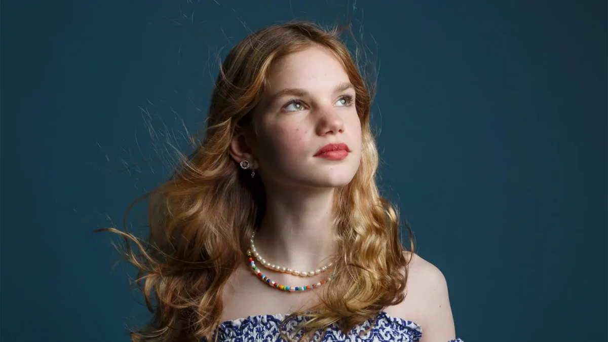 A young girl with curly hair, wearing a blue patterned top and adorned with a pearl necklace, gazes upwards thoughtfully against a deep blue background, embodying a mix of innocence and emerging elegance.